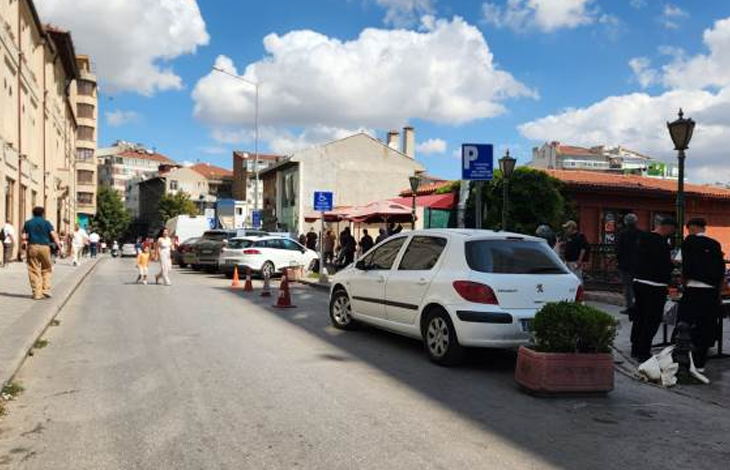 Görselin üst bölümünde, şehir merkezinde yer alan açık bir sokak ve otopark alanı yer alıyor. Yolun sağ kenarında, üzerinde tekerlekli sandalye simgesi bulunan mavi engelli park tabelasının hemen yanına usulsüz şekilde park etmiş beyaz renkli bir binek otomobil görülüyor; aracın önünde saksı içinde yeşil bir süs bitkisi, gerisinde ise duba ve koniler yer alıyor. Kaldırımda yürüyen yayalar, arka planda gölgelikli tezgâhlar ve dükkânlar ile güneşli, parçalı bulutlu gökyüzü altında uzanan şehir binaları seçiliyor.