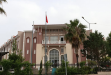 Görselin üst bölümünde, modern ve geleneksel Türk mimarisinden esintiler taşıyan Fethiye Adalet Sarayı'nın dış cephesi yer alıyor. Açık kahverengi ve krem tonlarındaki çok katlı binanın ön cephesinde, kemerli pencereler, beyaz korkuluklu bir balkon ve üzerinde büyük harflerle "FETHİYE ADALET SARAYI" yazısı görülüyor. Ön bahçede göndere çekilmiş Türk bayrağı, yüksek bir palmiye ağacı, yeşil ağaçlar ve demir korkuluklar bulunurken, üstte hafif bulutlu açık bir gökyüzü seçiliyor.