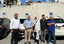 Görselin üst bölümünde, açık otopark alanında park edilmiş araçların önünde yan yana durarak poz veren beş kişilik bir vatandaş grubu yer alıyor. Soldan sağa doğru beyaz gömlekli ve siyah pantolonlu bir erkek, ekose kısa kollu gömlekli ve bej pantolonlu orta yaşlı bir erkek, beyaz başörtülü ve uzun beyaz tunikli bir kadın, siyah gömlekli genç bir erkek ve mavi polo tişörtlü kollarını kavuşturmuş yaşlıca bir adam bulunuyor. Grubun arkasındaki açık renkli yüksek duvarda mavi zeminli engelli park yeri tabelaları ("P" harfi ve tekerlekli sandalye sembolü) ile ortada beyaz bir Renault otomobil, sağda ise beyaz bir Fiat Egea araç görülüyor.