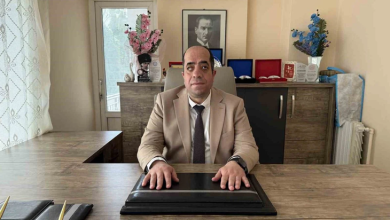 Geniş bir ahşap makam masasının arkasında oturan, açık kahverengi ceketli ve kravatlı görme engelli bir adam görülüyor. Elleri önündeki siyah sümen üzerinde duran kişinin arkasında ahşap konsol, üzerinde Atatürk portresi, çiçekler ve çeşitli plaketler yer alıyor.
