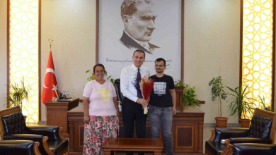 Görselin üst bölümünde, makam odasında gerçekleşen tebrik ziyaretinden neşeli bir an yer alıyor. Ortada beyaz gömlekli ve kravatlı rektör/yetkili, elinde kırmızı-mor renkli kuru çiçek buketini tutarak dururken; solunda açık pembe tişörtlü ve çiçek desenli etek giymiş tebessüm eden anne, sağında ise siyah baskılı tişört ve kot pantolon giymiş görme engelli üniversite öğrencisi Özcan Şaylı yer alıyor. Üçlü, arkalarında ahşap çalışma masası, büyük boy Mustafa Kemal Atatürk portresi ve Türk bayrağı ile yanlardaki deri koltukların bulunduğu resmi makam odasında yan yana kameraya gülümsüyor.