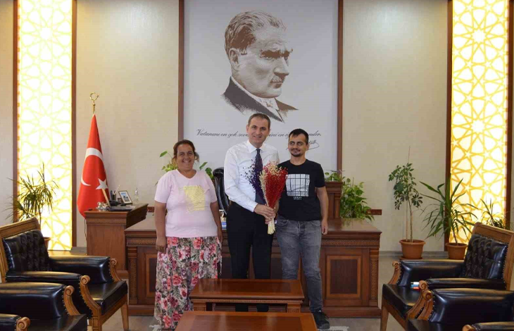 Görselin üst bölümünde, makam odasında gerçekleşen tebrik ziyaretinden neşeli bir an yer alıyor. Ortada beyaz gömlekli ve kravatlı rektör/yetkili, elinde kırmızı-mor renkli kuru çiçek buketini tutarak dururken; solunda açık pembe tişörtlü ve çiçek desenli etek giymiş tebessüm eden anne, sağında ise siyah baskılı tişört ve kot pantolon giymiş görme engelli üniversite öğrencisi Özcan Şaylı yer alıyor. Üçlü, arkalarında ahşap çalışma masası, büyük boy Mustafa Kemal Atatürk portresi ve Türk bayrağı ile yanlardaki deri koltukların bulunduğu resmi makam odasında yan yana kameraya gülümsüyor.