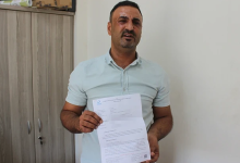Görselin üst bölümünde, sade bir oda ortamında ayakta durarak elindeki resmî evrakı kameraya doğru gösteren orta yaşlı bir adam yer alıyor. Kısa koyu renkli saçlı, hafif sakallı ve açık yeşil/mint tonlarında kısa kollu polo yaka tişört giymiş olan adam, yüzünde üzüntülü ve mağdur bir ifadeyle doğrudan objektife bakıyor. İki eliyle öne doğru tuttuğu A4 boyutundaki resmî belgenin sol üst köşesinde mavi renkli bir kurum logosu ve altında maddeler hâlinde yazılmış resmî metinler ile barkod detayı görülüyor; arka planda ise ahşap bir ofis dolabı ve beyaz duvar seçiliyor.