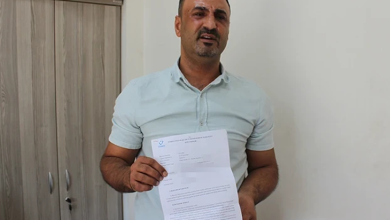 Görselin üst bölümünde, sade bir oda ortamında ayakta durarak elindeki resmî evrakı kameraya doğru gösteren orta yaşlı bir adam yer alıyor. Kısa koyu renkli saçlı, hafif sakallı ve açık yeşil/mint tonlarında kısa kollu polo yaka tişört giymiş olan adam, yüzünde üzüntülü ve mağdur bir ifadeyle doğrudan objektife bakıyor. İki eliyle öne doğru tuttuğu A4 boyutundaki resmî belgenin sol üst köşesinde mavi renkli bir kurum logosu ve altında maddeler hâlinde yazılmış resmî metinler ile barkod detayı görülüyor; arka planda ise ahşap bir ofis dolabı ve beyaz duvar seçiliyor.