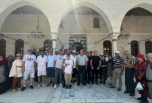 Görselin üst bölümünde, tarihi taş kemerli ve avlulu bir yapının önünde toplanmış, aralarında beyaz baston kullanan görme engelli bireylerin, refakatçilerinin ve çocukların bulunduğu kalabalık bir gezi grubu yer alıyor. Taş sütunlar ve kemerlerin altındaki avluda kadınlı erkekli yan yana dizilen grubun içinde sağ tarafta duran yaşlı bir bey beyaz bastonuna dayanırken, diğer katılımcılar yazlık kıyafetleriyle kameraya toplu hatıra pozu veriyor; arka planda tarihi yapının kemerleri, ferforje avizeleri ve ahşap detaylı pencereleri görülüyor.