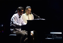 Görselin üst bölümünde, karanlık bir konser sahnesinde piyano başında yan yana performans sergileyen şarkıcı Karsu ile görme engelli müzisyen Şükrü Can Çapar yer alıyor. Sol tarafta piyano taburesinde oturan Çapar, siyah güneş gözlüğü, beyaz gömlek, siyah papyon ve kumaş pantolonuyla piyanonun tuşlarına basarak enstrümanı çalıyor. Sağ tarafında oturan Karsu ise sarı çiçek detaylı küpesi ve zarif beyaz sahne elbisesiyle piyanoya doğru eğilerek tebessümle mikrofon başında şarkı söylüyor; arka planda karanlık sahne atmosferi ve piyanonun yanında duran su şişesi ile nota sehpası görülüyor