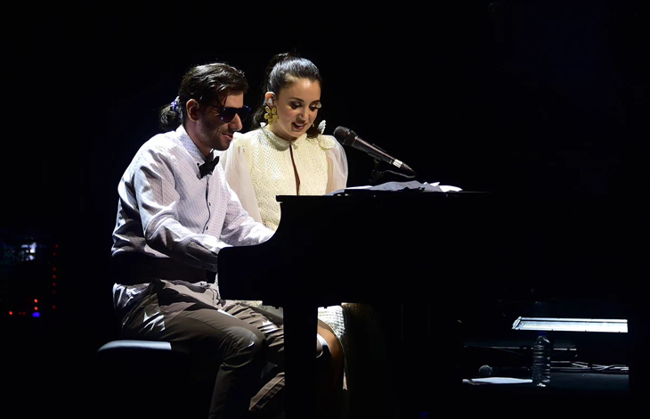 Görselin üst bölümünde, karanlık bir konser sahnesinde piyano başında yan yana performans sergileyen şarkıcı Karsu ile görme engelli müzisyen Şükrü Can Çapar yer alıyor. Sol tarafta piyano taburesinde oturan Çapar, siyah güneş gözlüğü, beyaz gömlek, siyah papyon ve kumaş pantolonuyla piyanonun tuşlarına basarak enstrümanı çalıyor. Sağ tarafında oturan Karsu ise sarı çiçek detaylı küpesi ve zarif beyaz sahne elbisesiyle piyanoya doğru eğilerek tebessümle mikrofon başında şarkı söylüyor; arka planda karanlık sahne atmosferi ve piyanonun yanında duran su şişesi ile nota sehpası görülüyor