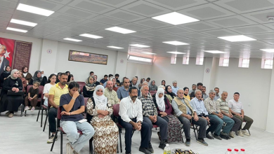 Görselde, geniş bir salonda sandalyelerde oturan kadın ve erkeklerden oluşan kalabalık bir grup görülüyor. Katılımcılar sıralar halinde otururken arka duvarda Türk bayrağı ve Atatürk görseli yer alıyor.