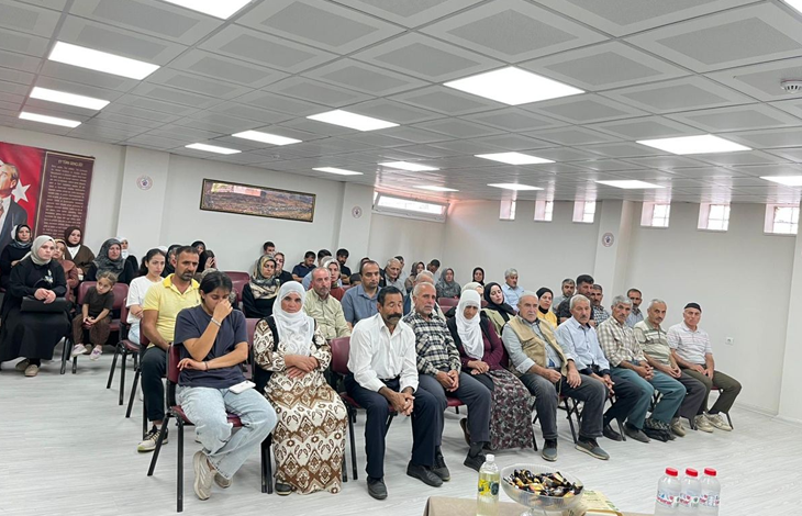 Görselde, geniş bir salonda sandalyelerde oturan kadın ve erkeklerden oluşan kalabalık bir grup görülüyor. Katılımcılar sıralar halinde otururken arka duvarda Türk bayrağı ve Atatürk görseli yer alıyor.