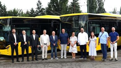 Açık havada, sarı renkli modern Anadolu Isuzu otobüslerinin bir sıra hâlinde park edildiği bir alan görülüyor. Otobüsler yeni, parlak ve kurumsal bir tanıtım etkinliği için hazırlanmış izlenimi veriyor. Ön kısımlarında marka logosu belirgin şekilde yer alıyor. Otobüslerin önünde, çoğu takım elbiseli ve resmi giyimli yaklaşık on iki kişilik bir yönetim ve temsilci grubu duruyor. Duruşları kurumsal bir tanıtım, teslim töreni veya basın etkinliği havası taşıyor. Arka planda uzun ağaçlar, elektrik hatları ve şehir yapıları bulunuyor. Genel atmosfer profesyonel, düzenli ve yeni araçların filoya katılımını simgeleyen bir tören görüntüsü sunuyor.
