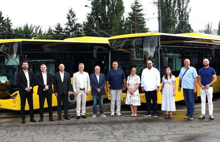 Açık havada, sarı renkli modern Anadolu Isuzu otobüslerinin bir sıra hâlinde park edildiği bir alan görülüyor. Otobüsler yeni, parlak ve kurumsal bir tanıtım etkinliği için hazırlanmış izlenimi veriyor. Ön kısımlarında marka logosu belirgin şekilde yer alıyor. Otobüslerin önünde, çoğu takım elbiseli ve resmi giyimli yaklaşık on iki kişilik bir yönetim ve temsilci grubu duruyor. Duruşları kurumsal bir tanıtım, teslim töreni veya basın etkinliği havası taşıyor. Arka planda uzun ağaçlar, elektrik hatları ve şehir yapıları bulunuyor. Genel atmosfer profesyonel, düzenli ve yeni araçların filoya katılımını simgeleyen bir tören görüntüsü sunuyor.