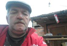 Instagram İçin Alternatif Metin: Görselin üst bölümünde, açık alanda selfie açısıyla çekilmiş portre fotoğrafında Mustafa Balkan yer alıyor. Gri renkli bir kasket takan, kır bıyıklı ve sakallı olan yaşlı adam, kırmızı bir mont ile altına örgü kazak giymiş vaziyette doğrudan kameraya bakıyor. Arka planda ise müstakil bir evin avlusu, ahşap sundurmalı ve tuğla duvarlı bir yapı ile çatı saçağından sarkan kırmızı detaylı bir temizlik fırçası görülüyor.