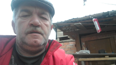 Instagram İçin Alternatif Metin: Görselin üst bölümünde, açık alanda selfie açısıyla çekilmiş portre fotoğrafında Mustafa Balkan yer alıyor. Gri renkli bir kasket takan, kır bıyıklı ve sakallı olan yaşlı adam, kırmızı bir mont ile altına örgü kazak giymiş vaziyette doğrudan kameraya bakıyor. Arka planda ise müstakil bir evin avlusu, ahşap sundurmalı ve tuğla duvarlı bir yapı ile çatı saçağından sarkan kırmızı detaylı bir temizlik fırçası görülüyor.