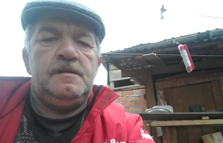 Instagram İçin Alternatif Metin: Görselin üst bölümünde, açık alanda selfie açısıyla çekilmiş portre fotoğrafında Mustafa Balkan yer alıyor. Gri renkli bir kasket takan, kır bıyıklı ve sakallı olan yaşlı adam, kırmızı bir mont ile altına örgü kazak giymiş vaziyette doğrudan kameraya bakıyor. Arka planda ise müstakil bir evin avlusu, ahşap sundurmalı ve tuğla duvarlı bir yapı ile çatı saçağından sarkan kırmızı detaylı bir temizlik fırçası görülüyor.
