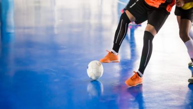 Görselde, kapalı bir sahada turuncu formalı bir futbolcunun top sürerken, rakibinin ise onu takip ettiği bir futsal maçı görülüyor.