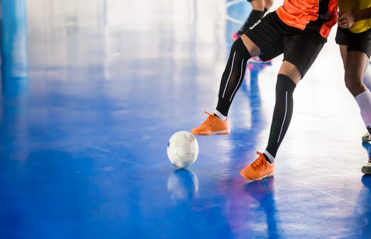 Görselde, kapalı bir sahada turuncu formalı bir futbolcunun top sürerken, rakibinin ise onu takip ettiği bir futsal maçı görülüyor.