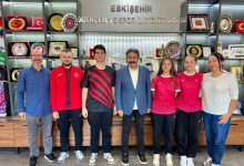 Instagram İçin Alternatif Metin:Görselin üst bölümünde, Eskişehir Gençlik ve Spor İl Müdürlüğü makam odasında yan yana dizilerek poz veren milli sporcular, antrenörler ve il müdürü yer alıyor. Arka plandaki duvarda gümüş renkli kabartma harflerle "ESKİŞEHİR GENÇLİK VE SPOR İL MÜDÜRLÜĞÜ" yazısı ile plaketler, kupalar ve tabaklarla dolu vitrinli ahşap bir sergi dolabı görülüyor. Mavi ceketli ve beyaz gömlekli müdürün sağında ve solunda; kırmızı milli takım fermuarlı üstü ve siyah antrenman formaları giymiş sporcular ile kırmızı formalarıyla işitme engelli kadın milli badmintoncular yer alıyor. Grubun en solunda mavi gömlekli, gözlüklü antrenör; en sağında ise beyaz bluzlu ve kot pantolonlu bir kadın yetkili durarak hep birlikte objektife gülümsüyor.
