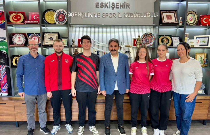 Instagram İçin Alternatif Metin:Görselin üst bölümünde, Eskişehir Gençlik ve Spor İl Müdürlüğü makam odasında yan yana dizilerek poz veren milli sporcular, antrenörler ve il müdürü yer alıyor. Arka plandaki duvarda gümüş renkli kabartma harflerle "ESKİŞEHİR GENÇLİK VE SPOR İL MÜDÜRLÜĞÜ" yazısı ile plaketler, kupalar ve tabaklarla dolu vitrinli ahşap bir sergi dolabı görülüyor. Mavi ceketli ve beyaz gömlekli müdürün sağında ve solunda; kırmızı milli takım fermuarlı üstü ve siyah antrenman formaları giymiş sporcular ile kırmızı formalarıyla işitme engelli kadın milli badmintoncular yer alıyor. Grubun en solunda mavi gömlekli, gözlüklü antrenör; en sağında ise beyaz bluzlu ve kot pantolonlu bir kadın yetkili durarak hep birlikte objektife gülümsüyor.