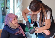 Görselin üst bölümünde, ev ortamında bir yaşlı kadının tansiyonunu ölçen belediye evde sağlık hizmeti görevlisi yer alıyor. Sol tarafta sandalyede oturan pembe-mor desenli başörtülü yaşlı kadın tebessümle sağlık görevlisine bakarken; sağ tarafta siyah eldivenli, yelekli ve beyaz tişörtlü kadın sağlık personeli özenle eğilerek kadının koluna yeşil renkli tansiyon aletinin manşonunu bağlıyor. Arka planda pencere, beyaz tül perde ve sehpa üzerindeki su şişesi seçiliyor.
