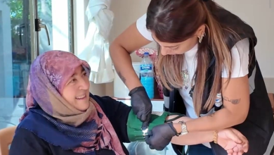 Görselin üst bölümünde, ev ortamında bir yaşlı kadının tansiyonunu ölçen belediye evde sağlık hizmeti görevlisi yer alıyor. Sol tarafta sandalyede oturan pembe-mor desenli başörtülü yaşlı kadın tebessümle sağlık görevlisine bakarken; sağ tarafta siyah eldivenli, yelekli ve beyaz tişörtlü kadın sağlık personeli özenle eğilerek kadının koluna yeşil renkli tansiyon aletinin manşonunu bağlıyor. Arka planda pencere, beyaz tül perde ve sehpa üzerindeki su şişesi seçiliyor.