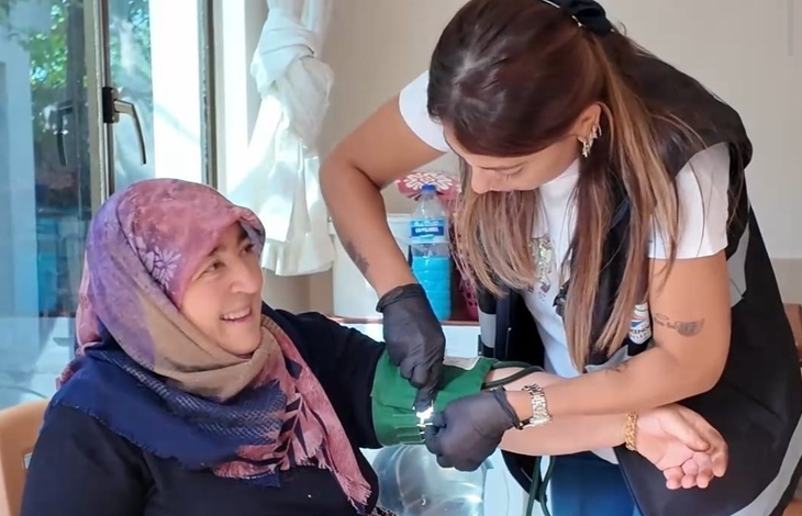 Görselin üst bölümünde, ev ortamında bir yaşlı kadının tansiyonunu ölçen belediye evde sağlık hizmeti görevlisi yer alıyor. Sol tarafta sandalyede oturan pembe-mor desenli başörtülü yaşlı kadın tebessümle sağlık görevlisine bakarken; sağ tarafta siyah eldivenli, yelekli ve beyaz tişörtlü kadın sağlık personeli özenle eğilerek kadının koluna yeşil renkli tansiyon aletinin manşonunu bağlıyor. Arka planda pencere, beyaz tül perde ve sehpa üzerindeki su şişesi seçiliyor.