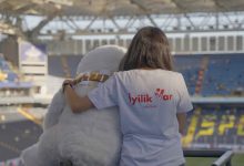 Bir stadyumda, sırtı kameraya dönük bir kişi görülüyor. Üzerinde beyaz bir tişört var ve tişörtün arka kısmında kırmızı renkle “İyilik Var” ve “#İyilikVar” yazıyor. Kişi, yanında oturan büyük, beyaz, peluş görünümlü bir maskotun omzuna kolunu dolamış. Maskot sakin ve sevimli bir duruş sergiliyor. Arka planda mavi ve sarı koltukların bulunduğu geniş bir tribün ve üstü kapalı stadyum çatısı görünüyor. Sahne, sıcaklık ve dayanışma hissi veriyor.