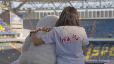 Bir stadyumda, sırtı kameraya dönük bir kişi görülüyor. Üzerinde beyaz bir tişört var ve tişörtün arka kısmında kırmızı renkle “İyilik Var” ve “#İyilikVar” yazıyor. Kişi, yanında oturan büyük, beyaz, peluş görünümlü bir maskotun omzuna kolunu dolamış. Maskot sakin ve sevimli bir duruş sergiliyor. Arka planda mavi ve sarı koltukların bulunduğu geniş bir tribün ve üstü kapalı stadyum çatısı görünüyor. Sahne, sıcaklık ve dayanışma hissi veriyor.