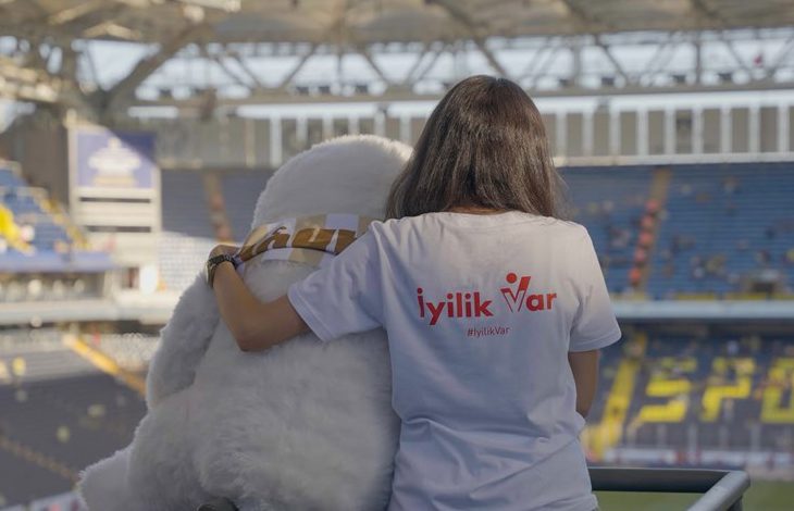 Bir stadyumda, sırtı kameraya dönük bir kişi görülüyor. Üzerinde beyaz bir tişört var ve tişörtün arka kısmında kırmızı renkle “İyilik Var” ve “#İyilikVar” yazıyor. Kişi, yanında oturan büyük, beyaz, peluş görünümlü bir maskotun omzuna kolunu dolamış. Maskot sakin ve sevimli bir duruş sergiliyor. Arka planda mavi ve sarı koltukların bulunduğu geniş bir tribün ve üstü kapalı stadyum çatısı görünüyor. Sahne, sıcaklık ve dayanışma hissi veriyor.