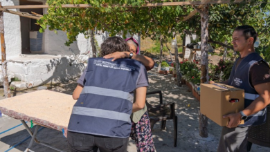 Görselin üst bölümünde, kırsal veya müstakil bir evin bahçesinde gerçekleştirilen sosyal destek ziyaretinden samimi bir an yer alıyor. Üzerinde "İZMİR BÜYÜKŞEHİR BELEDİYESİ SOSYAL HİZMETLER DAİRESİ BAŞKANLIĞI" yazılı lacivert yelek giymiş bir sosyal hizmet görevlisi, yaşlı bir kadınla sarılarak kucaklaşırken; sağ tarafta yine aynı belediye yeleğini giyen başka bir görevli elinde yardım kolisiyle onları gülümseyerek izliyor. Bahçede üstü asmalarla örtülü çardak, ahşap bir masa ve arka planda köy evi detayları görülüyor.