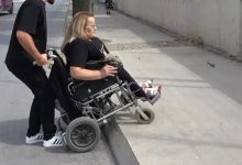 Bir kişi, rampa olmadığı için tekerlekli sandalyesiyle yüksek kaldırıma çıkmaya çalışıyor. Sandalyede oturan kadının arkasındaki erkek, sandalyenin ön tarafını havaya kaldırarak onu kaldırımın üzerine çıkarmaya yardımcı oluyor. Sandalyenin ön tekerlekleri kaldırımın aşağısına uzanmışken arka tekerlekler yukarıda duruyor. Sahne dış mekânda, beton bir yol üzerinde geçiyor.