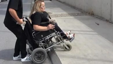 Bir kişi, rampa olmadığı için tekerlekli sandalyesiyle yüksek kaldırıma çıkmaya çalışıyor. Sandalyede oturan kadının arkasındaki erkek, sandalyenin ön tarafını havaya kaldırarak onu kaldırımın üzerine çıkarmaya yardımcı oluyor. Sandalyenin ön tekerlekleri kaldırımın aşağısına uzanmışken arka tekerlekler yukarıda duruyor. Sahne dış mekânda, beton bir yol üzerinde geçiyor.