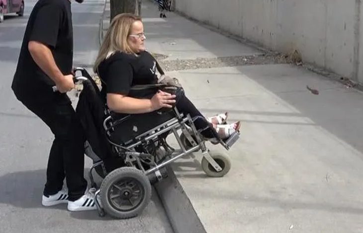 Bir kişi, rampa olmadığı için tekerlekli sandalyesiyle yüksek kaldırıma çıkmaya çalışıyor. Sandalyede oturan kadının arkasındaki erkek, sandalyenin ön tarafını havaya kaldırarak onu kaldırımın üzerine çıkarmaya yardımcı oluyor. Sandalyenin ön tekerlekleri kaldırımın aşağısına uzanmışken arka tekerlekler yukarıda duruyor. Sahne dış mekânda, beton bir yol üzerinde geçiyor.