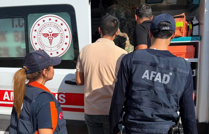 Görselin üst bölümünde, beyaz renkli bir ambulansın açık arka kapısı önünde toplanmış arama kurtarma personeli, sağlık görevlileri ve bulunan genç adam yer alıyor. Ön planda sırtında büyük beyaz harflerle "AFAD" yazılı lacivert yelek giymiş ve lacivert AFAD şapkası takmış bir görevli ile sol tarafında at kuyruklu saçları olan kadın bir sağlık/kurtarma personeli duruyor. Ortada ise bej renkli kısa kollu polo tişört giymiş genç adam, sağlık kontrolü ve ilk müdahale için ambulansa doğru yönlendirilirken görülüyor; arka planda ambulansın iç donanımı ve araç üzerindeki kırmızı-beyaz T.C. Sağlık Bakanlığı amblemi dikkat çekiyor.