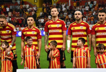 Kırmızı–sarı Kayserispor forması giyen futbolcular, maç öncesi sahada yan yana duruyor. Önlerinde aynı renklerde forma giymiş down sendromlu çocuklar bulunuyor. Tribünler dolu, atmosfer sıcak ve destekleyici.