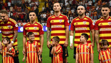 Kırmızı–sarı Kayserispor forması giyen futbolcular, maç öncesi sahada yan yana duruyor. Önlerinde aynı renklerde forma giymiş down sendromlu çocuklar bulunuyor. Tribünler dolu, atmosfer sıcak ve destekleyici.