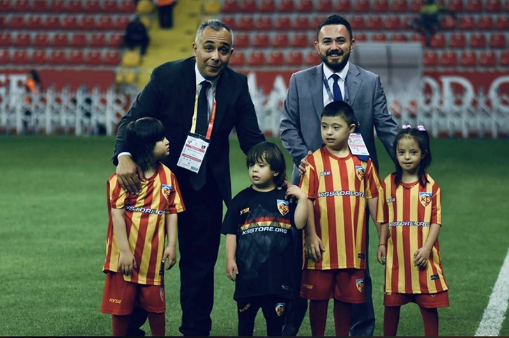 Kayserispor yöneticileri saha kenarında ayakta duruyor. Yanlarında kırmızı–sarı formalar giymiş down sendromlu çocuklar bulunuyor. Arka planda tribünler ve reklam panoları görülüyor. Görsel, kulüp yöneticileri ile çocukların birlikte yer aldığı samimi bir anı yansıtıyor.