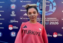 Para Yüzücü Sevilay Öztürk üstünde Türkiye yazılı mercan rengi tişörtle turnuva panosu önünde poz veriyor.