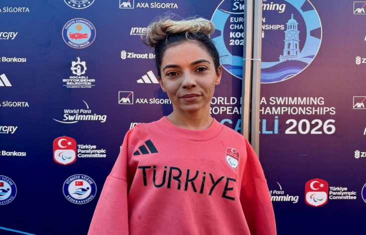 Para Yüzücü Sevilay Öztürk üstünde Türkiye yazılı mercan rengi tişörtle turnuva panosu önünde poz veriyor.