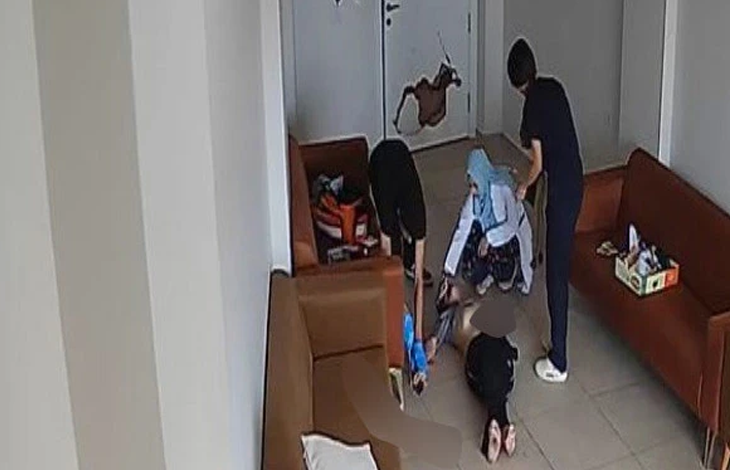 Görselin üst bölümünde, bir rehabilitasyon merkezinin bekleme veya dinlenme odasını yukarıdan gören güvenlik kamerası görüntüsü yer alıyor. Kahverengi deri koltukların bulunduğu odanın zemininde sırtüstü hareketsiz yatan bir birey, başında diz çökmüş mavi başörtülü bir kadın görevli ve ayakta duran personeller görülüyor. Arka plandaki beyaz kapının üzerinde büyük bir delik ve kırık hasarı dikkat çekerken, görevlilerin yerde yatan bireye müdahale ettiği anlar yansıyor.