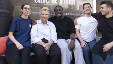 Görselin üst bölümünde, bir "Cafe & Fırın" tabelasının önünde yan yana oturarak samimi bir şekilde poz veren beş kişilik neşeli bir grup yer alıyor. Soldan sağa doğru gözlüklü mavi tişörtlü bir genç kadın, ortada beyaz gömlekli yaşlı bir kadın, onun yanında siyah tişörtlü gülümseyen siyahi bir genç, gözlüklü beyaz tişörtlü Down sendromlu bir genç ve en sağda siyah tişörtlü başka bir genç çalışan yan yana oturuyor. Arkadaki cam vitrin üzerinde "Cafe & Fırın" yazısı ve bilgilendirici ibareler seçiliyor.