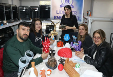 Bir atölye ortamında, beş kişi geniş bir masanın etrafında oturuyor. Masanın üzerinde renkli 3D yazıcı çıktıları bulunuyor; geometrik şekiller, küçük figürler, dekoratif objeler ve çeşitli tasarım örnekleri dikkat çekiyor. Arkada raflara yerleştirilmiş birden fazla 3D yazıcı görülüyor. Duvar üzerindeki “Liderler Enstitüsü” yazılı afiş, ortamın eğitim ve teknoloji odaklı bir çalışma alanı olduğunu gösteriyor.