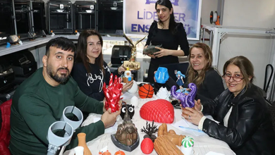 Bir atölye ortamında, beş kişi geniş bir masanın etrafında oturuyor. Masanın üzerinde renkli 3D yazıcı çıktıları bulunuyor; geometrik şekiller, küçük figürler, dekoratif objeler ve çeşitli tasarım örnekleri dikkat çekiyor. Arkada raflara yerleştirilmiş birden fazla 3D yazıcı görülüyor. Duvar üzerindeki “Liderler Enstitüsü” yazılı afiş, ortamın eğitim ve teknoloji odaklı bir çalışma alanı olduğunu gösteriyor.