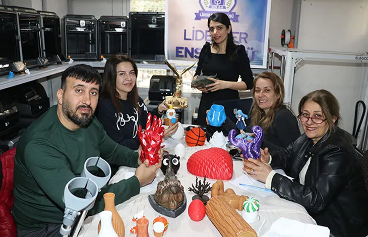 Bir atölye ortamında, beş kişi geniş bir masanın etrafında oturuyor. Masanın üzerinde renkli 3D yazıcı çıktıları bulunuyor; geometrik şekiller, küçük figürler, dekoratif objeler ve çeşitli tasarım örnekleri dikkat çekiyor. Arkada raflara yerleştirilmiş birden fazla 3D yazıcı görülüyor. Duvar üzerindeki “Liderler Enstitüsü” yazılı afiş, ortamın eğitim ve teknoloji odaklı bir çalışma alanı olduğunu gösteriyor.