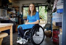 Lucy Robinson, tekerlekli sandalyesinde oturmuş şekilde aydınlık bir çalışma alanında görülüyor. Üzerinde mavi bir elbise, koyu renk tayt ve renkli bağcıklı beyaz spor ayakkabılar var. Arka planda raflar, kutular, bir çalışma masası ve cam kapıdan görünen bitkilerle sakin, düzenli bir ortam dikkat çekiyor.