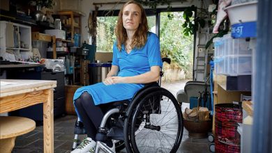 Lucy Robinson, tekerlekli sandalyesinde oturmuş şekilde aydınlık bir çalışma alanında görülüyor. Üzerinde mavi bir elbise, koyu renk tayt ve renkli bağcıklı beyaz spor ayakkabılar var. Arka planda raflar, kutular, bir çalışma masası ve cam kapıdan görünen bitkilerle sakin, düzenli bir ortam dikkat çekiyor.
