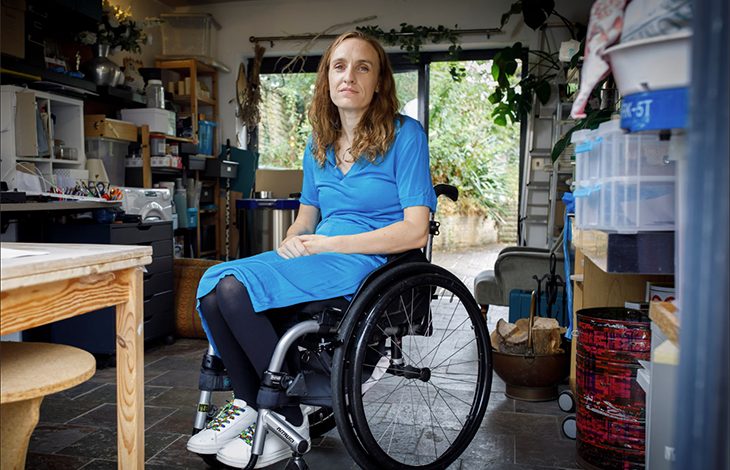 Lucy Robinson, tekerlekli sandalyesinde oturmuş şekilde aydınlık bir çalışma alanında görülüyor. Üzerinde mavi bir elbise, koyu renk tayt ve renkli bağcıklı beyaz spor ayakkabılar var. Arka planda raflar, kutular, bir çalışma masası ve cam kapıdan görünen bitkilerle sakin, düzenli bir ortam dikkat çekiyor.