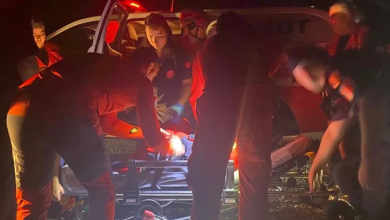 Görselin üst bölümünde, gece karanlığında acil servis veya arama kurtarma aracının tepe lambalarının kırmızı ışığı altında sedye etrafında toplanmış sağlık ve kurtarma ekipleri yer alıyor. Kırmızı üniformalı ve fosfor detaylı kıyafetler giymiş acil müdahale personeli, bulunan vatandaşı taşımak üzere hazırlanan tekerlekli ambulans sedyesinin çevresinde eğilmiş vaziyette ilk müdahaleyi ve sabitlemeyi gerçekleştiriyor; arka planda açık kapılı beyaz bir kurtarma aracı ve ekiplerin dikkatli çalışmaları görülüyor.