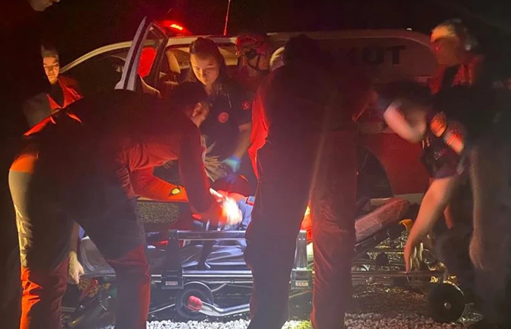 Görselin üst bölümünde, gece karanlığında acil servis veya arama kurtarma aracının tepe lambalarının kırmızı ışığı altında sedye etrafında toplanmış sağlık ve kurtarma ekipleri yer alıyor. Kırmızı üniformalı ve fosfor detaylı kıyafetler giymiş acil müdahale personeli, bulunan vatandaşı taşımak üzere hazırlanan tekerlekli ambulans sedyesinin çevresinde eğilmiş vaziyette ilk müdahaleyi ve sabitlemeyi gerçekleştiriyor; arka planda açık kapılı beyaz bir kurtarma aracı ve ekiplerin dikkatli çalışmaları görülüyor.