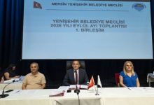 Görselde, resmi bir toplantı salonunda sahne üzerindeki masada yan yana oturan üç kişi görülüyor. Masanın üzerinde mikrofonlar, belgeler ve küçük bayraklar bulunuyor. Arkadaki büyük ekranda belediye meclisi toplantısına ait başlık yer alıyor. Ortam ciddi, düzenli ve kurumsal bir meclis oturumu atmosferi taşıyor.