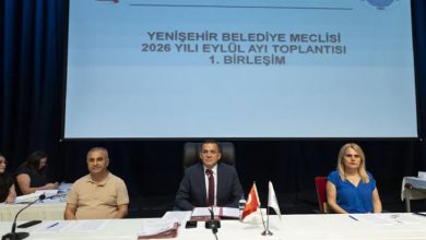Görselde, resmi bir toplantı salonunda sahne üzerindeki masada yan yana oturan üç kişi görülüyor. Masanın üzerinde mikrofonlar, belgeler ve küçük bayraklar bulunuyor. Arkadaki büyük ekranda belediye meclisi toplantısına ait başlık yer alıyor. Ortam ciddi, düzenli ve kurumsal bir meclis oturumu atmosferi taşıyor.